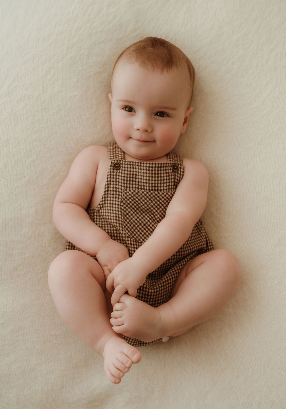 Baby wearing brown gingham romper sits on cream colored background in studio portrait setting.