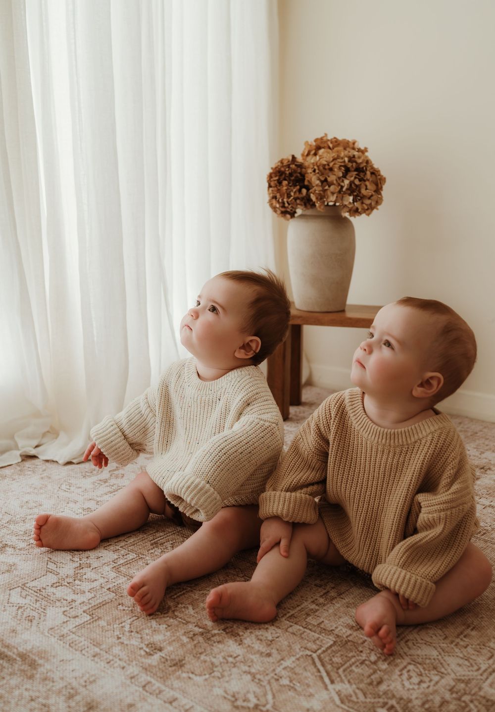 Two babies in matching beige knit sweaters lay on a floral blanket looking upward with playful expressions.