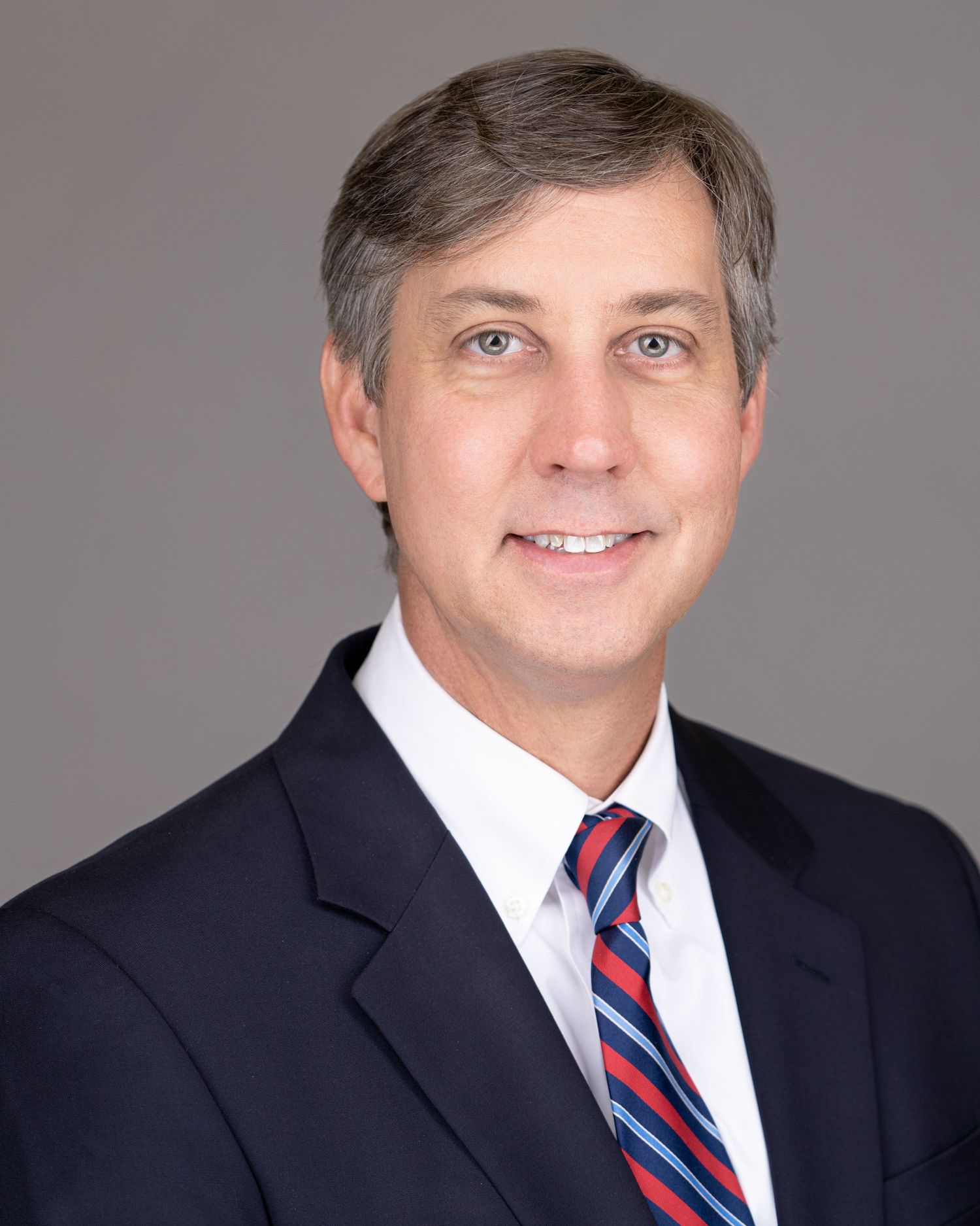 Professional headshot portrait of  a man wearing a dark suit and red striped tie against a gray background in Charleston, SC