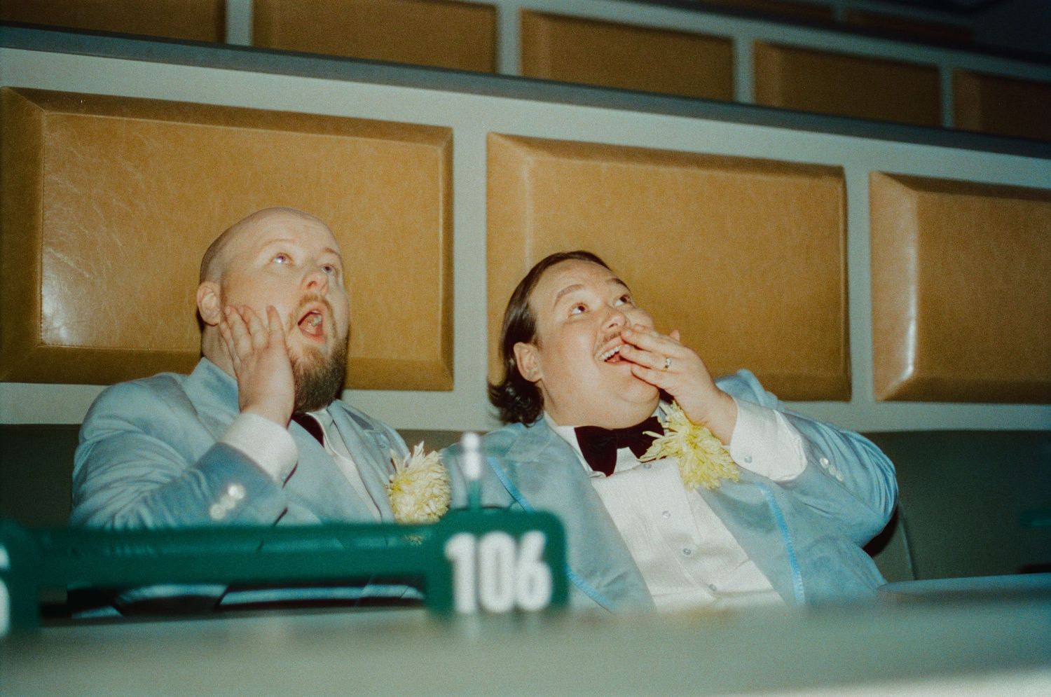 Two men in light blue suits sit in stadium seats, eating popcorn and looking upward with surprised expressions.