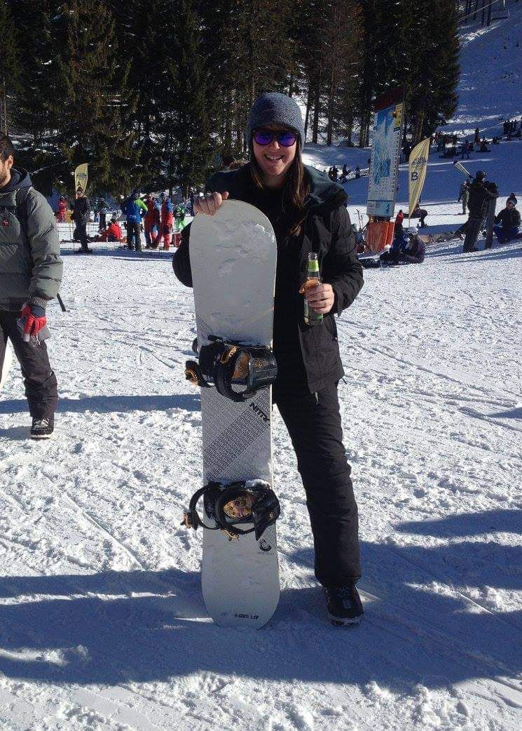 Person holding a snowboard on snowy slopes with other winter sports enthusiasts visible.