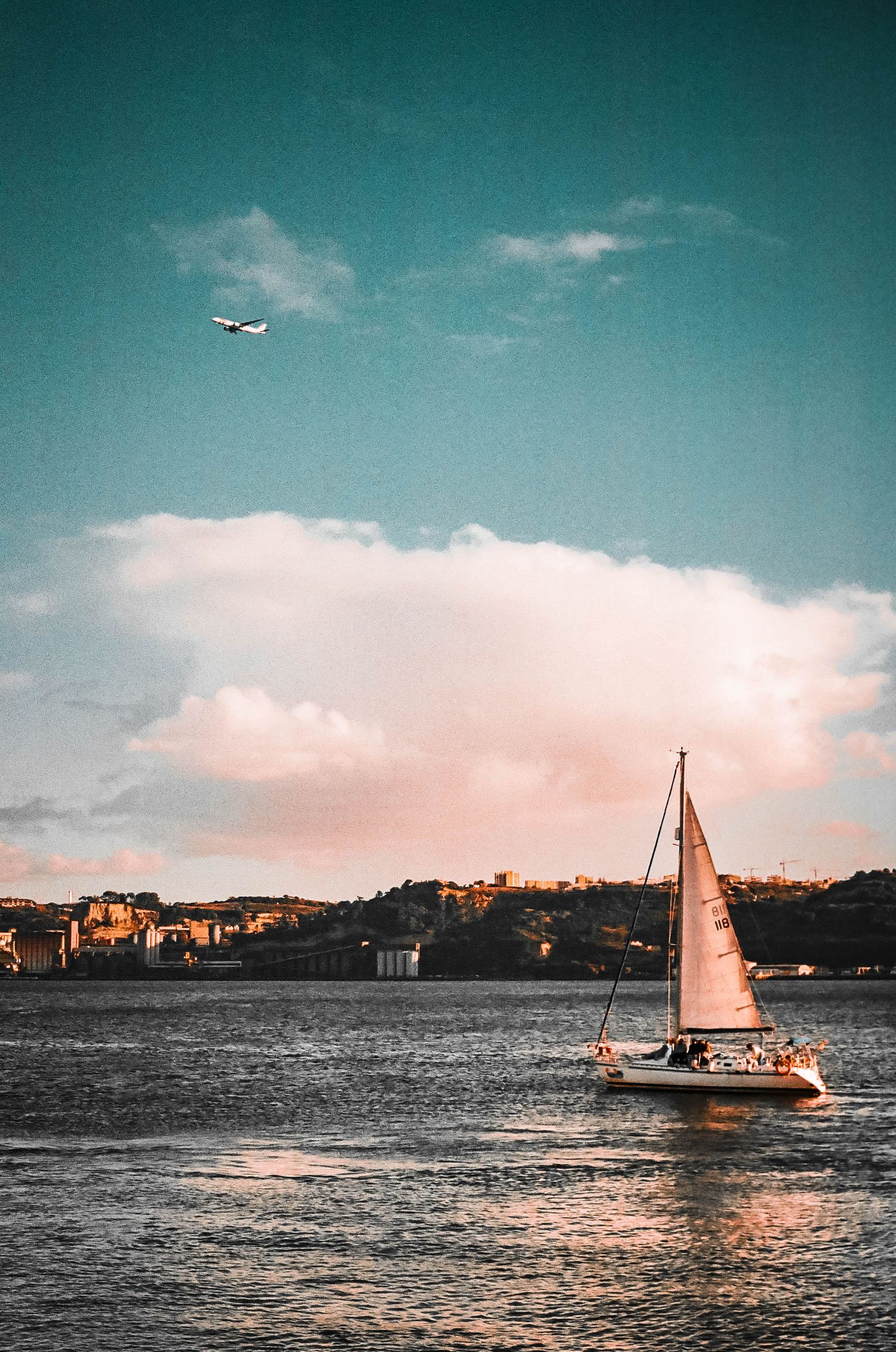 Sailboat cruising on water at sunset with pink clouds and airplane in teal sky.