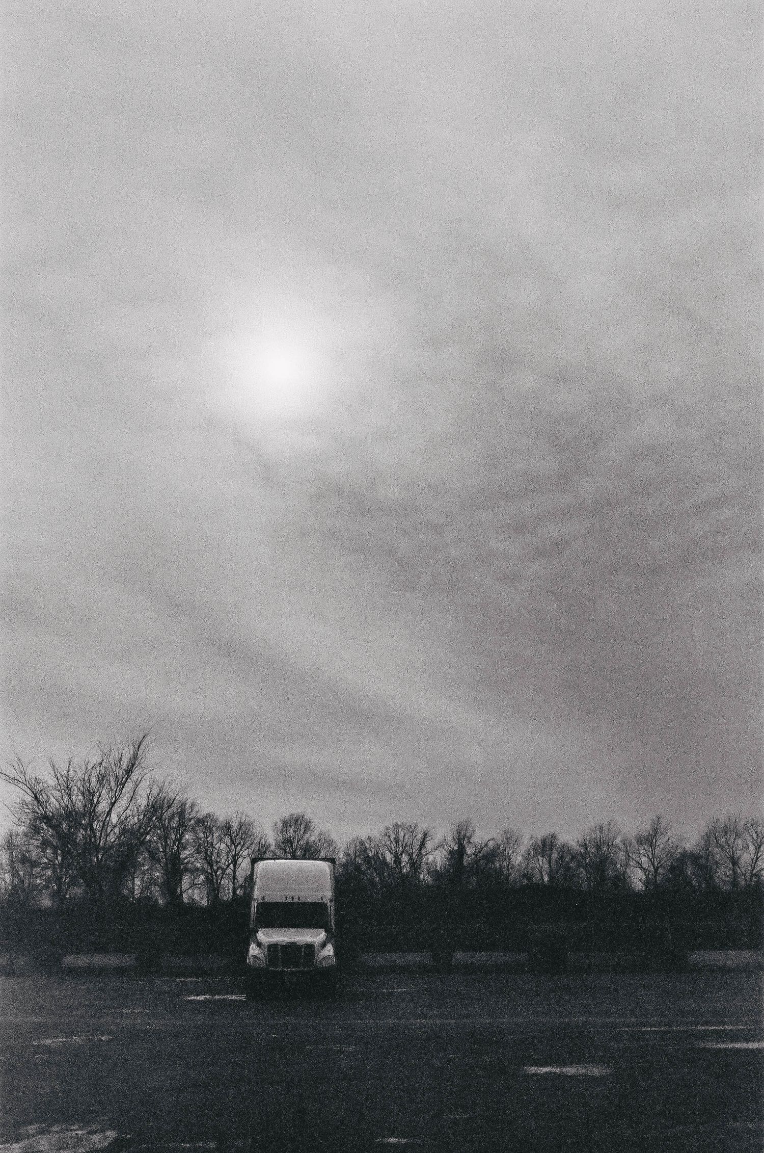 A solitary van parked under a moody overcast sky with sun peeking through clouds and silhouetted trees in background.