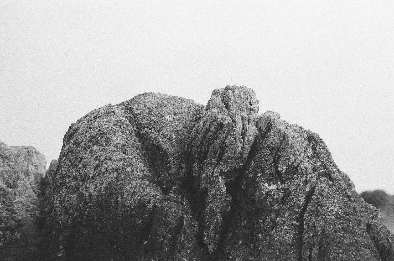A black and white photograph of a rugged mountain peak with jagged rock formations against a light sky.