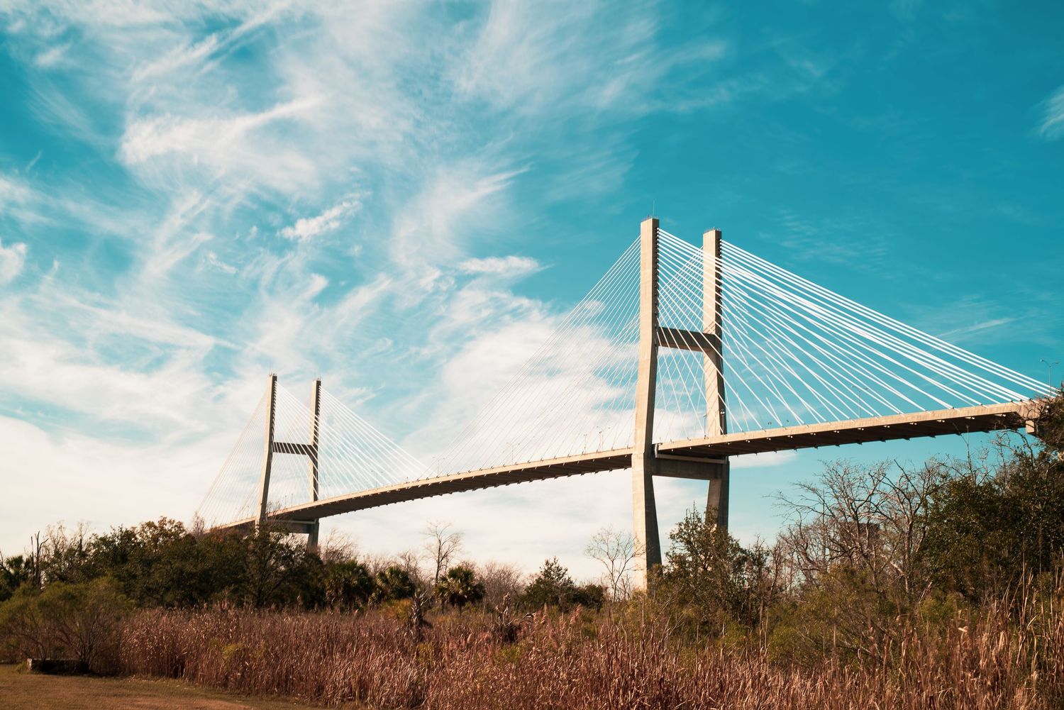 Cable-stayed bridge spans across water against vibrant turquoise sky with golden sunlight illuminating its structure.
