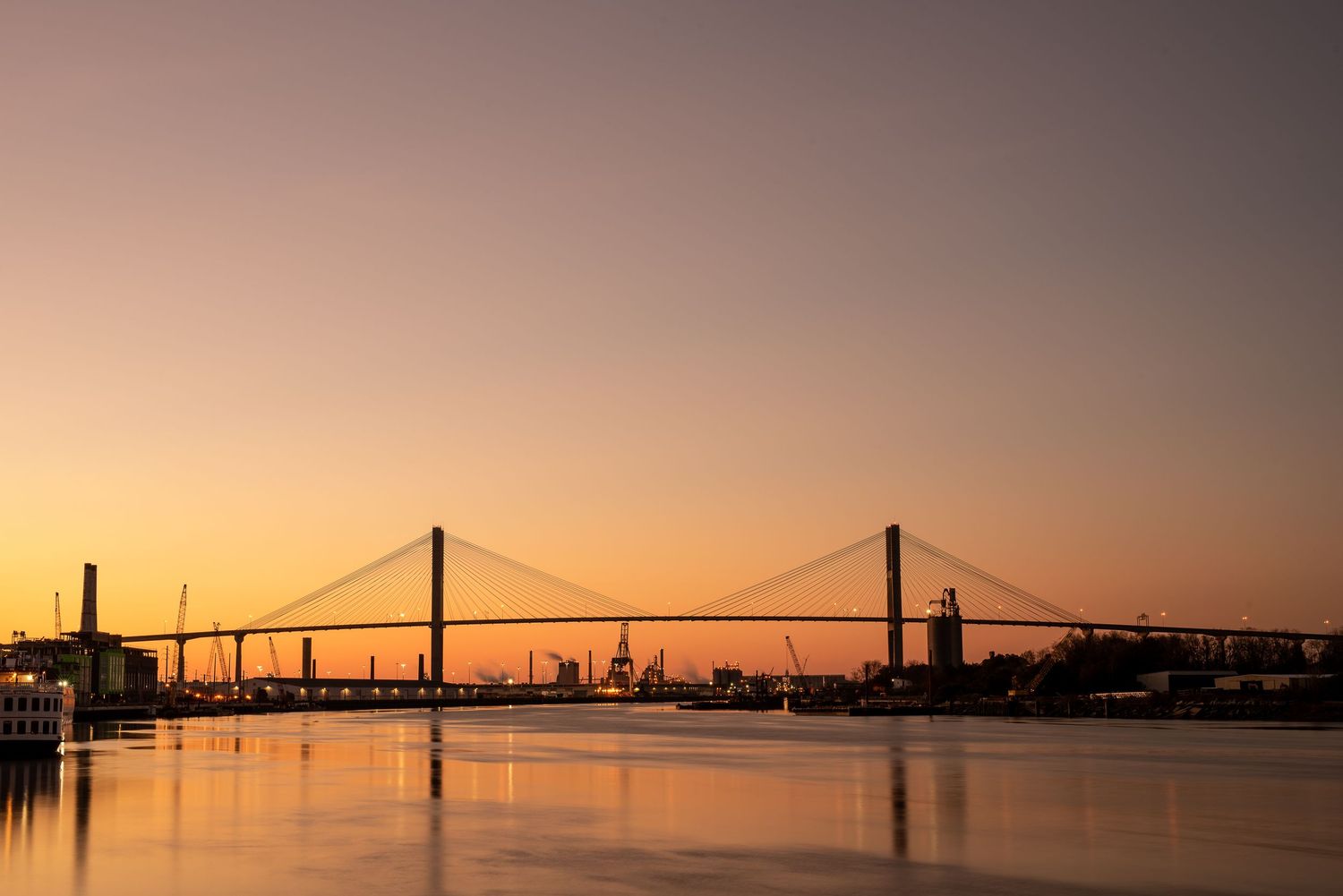 Panoramic silhouette of suspension bridge and city skyline at sunset over calm water.