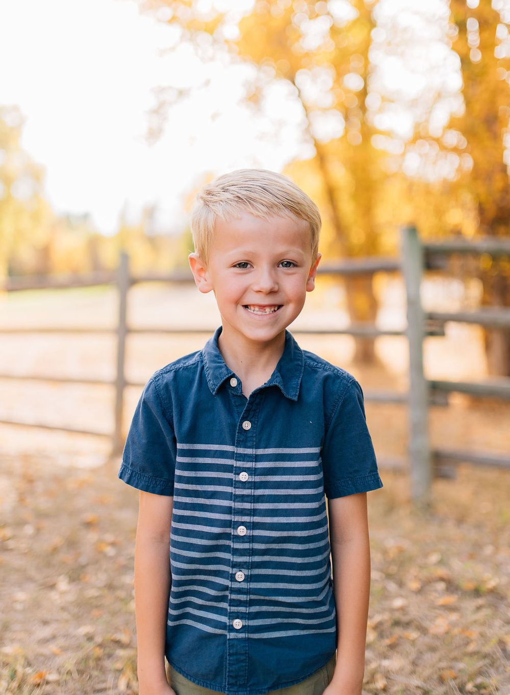 A young child wearing a blue and white striped button-down shirt stands outdoors with autumn trees and a wooden fence in background.