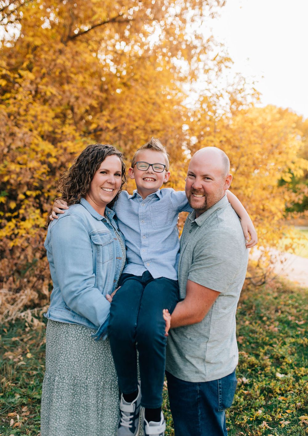 A family poses together outdoors against golden autumn foliage on a sunny fall day.