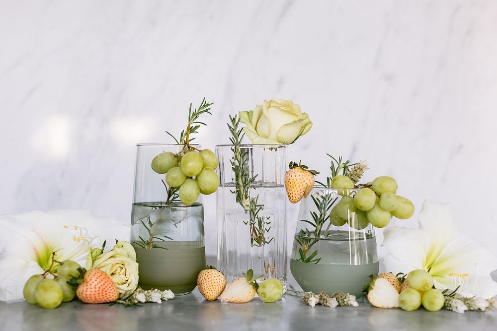 Green cocktails garnished with rosemary and white roses surrounded by grapes on marble surface.