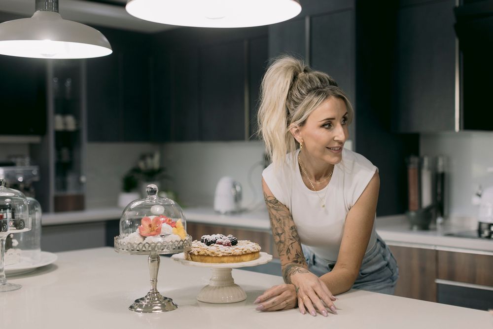 Tattooed blonde woman smiling in modern kitchen with decorated cakes on elegant cake stands.