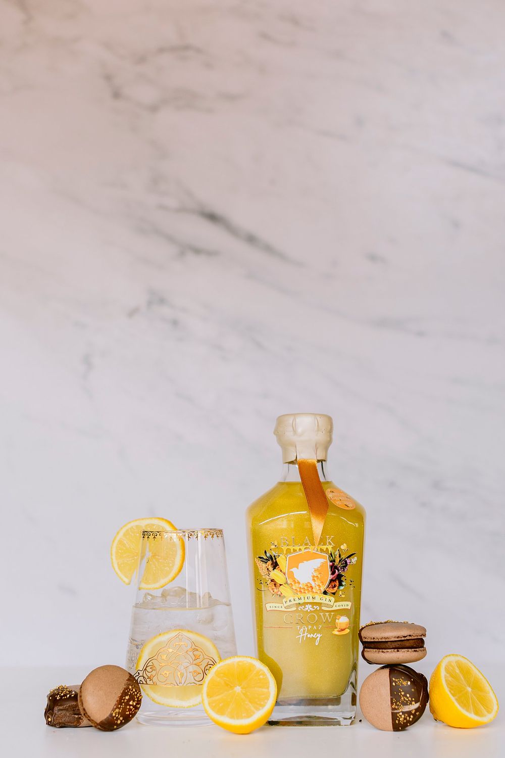 Yellow honey gin bottle with lemon slices, chocolate macarons, and a cocktail glass on marble.