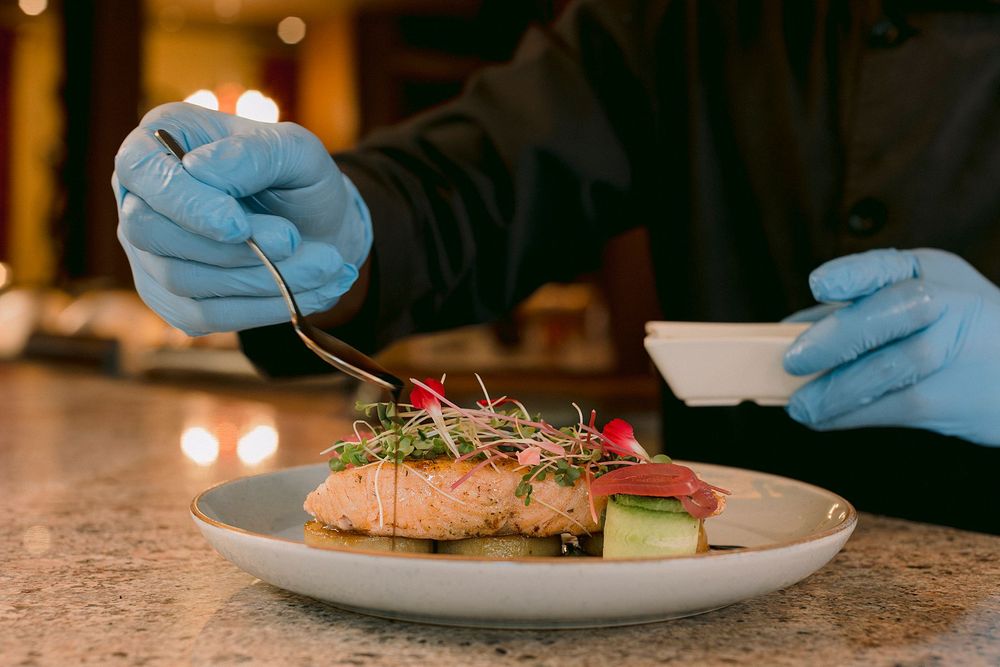 Chef in blue gloves plating fresh salmon dish with microgreens and vegetables on white plate.
