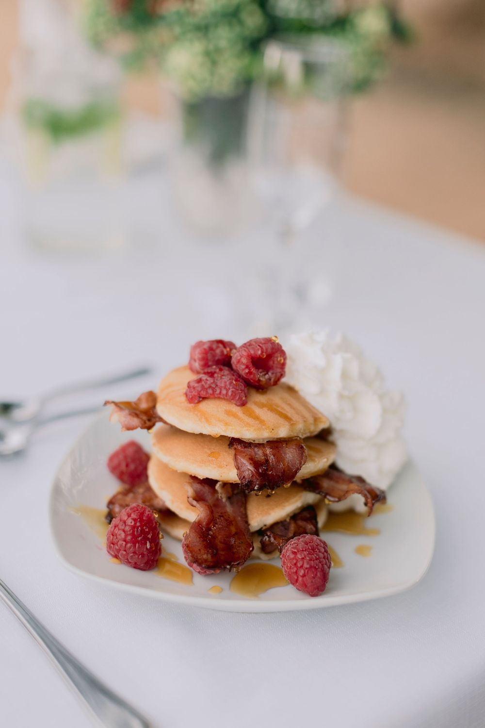 Stacked pancakes with fresh raspberries, crispy bacon, whipped cream, and maple syrup on plate.