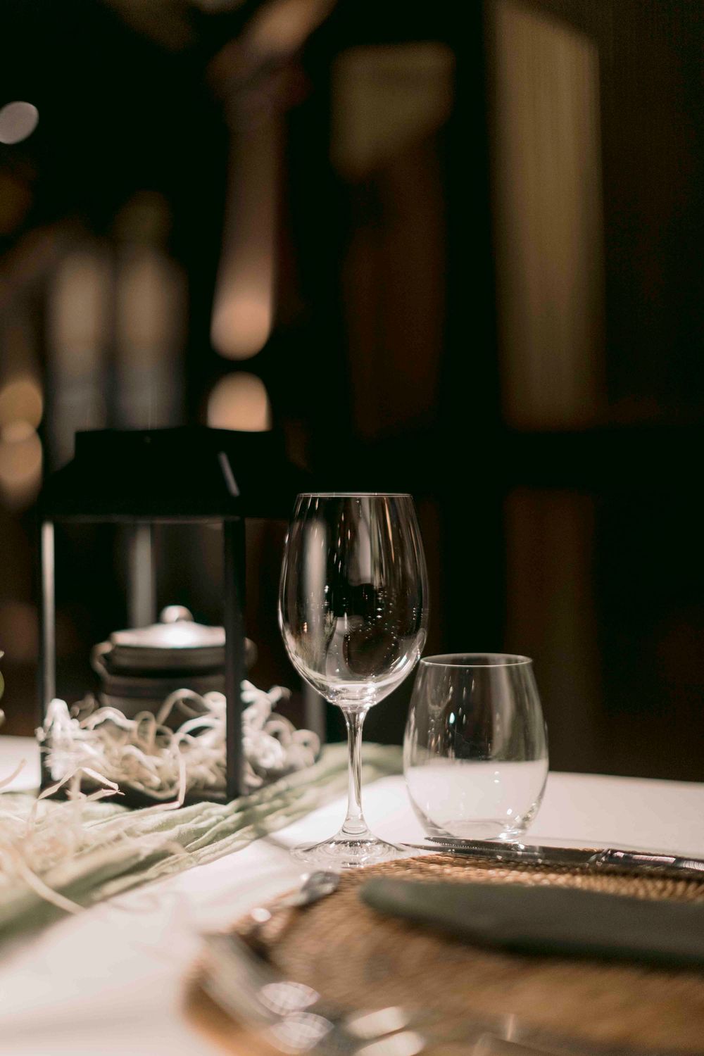 Close-up of empty wine glass and tumbler on a white tablecloth with a dark lantern centerpiece in a restaurant.