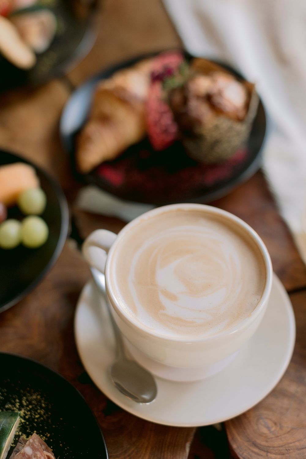 Close-up of creamy latte art in white cup with blurred breakfast pastries and fruit in background.
