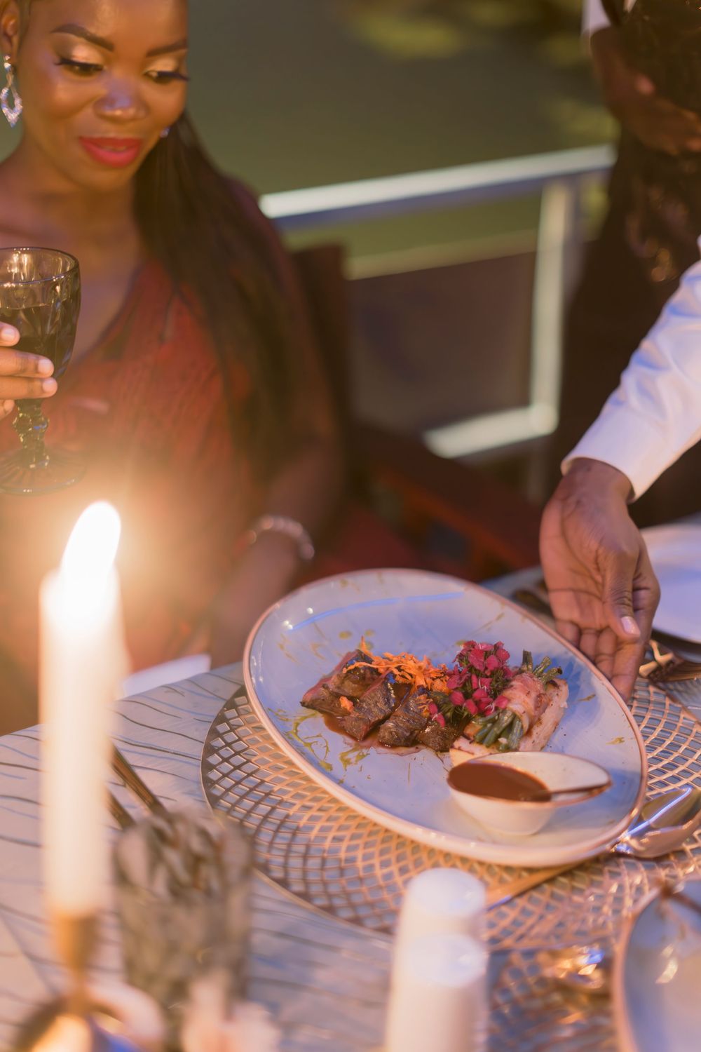 Waiter presenting a gourmet steak dish to a woman holding wine at a romantic candlelit dinner.