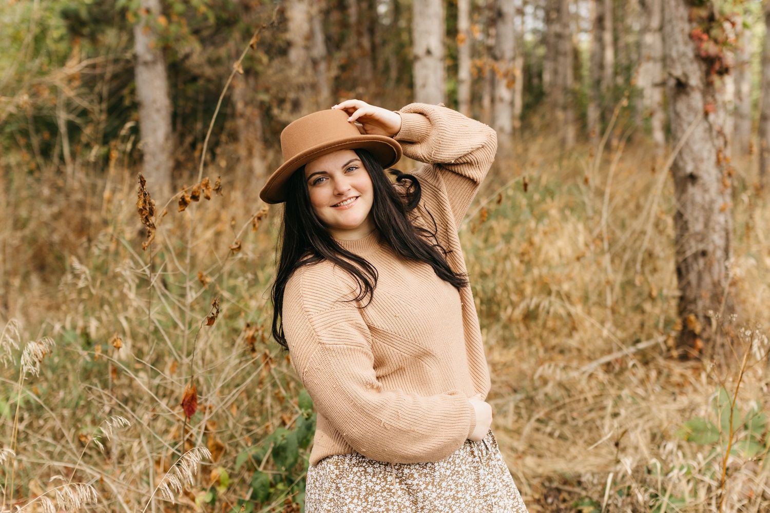 A person in a brown dress and hat poses thoughtfully in a sunlit autumn forest setting.