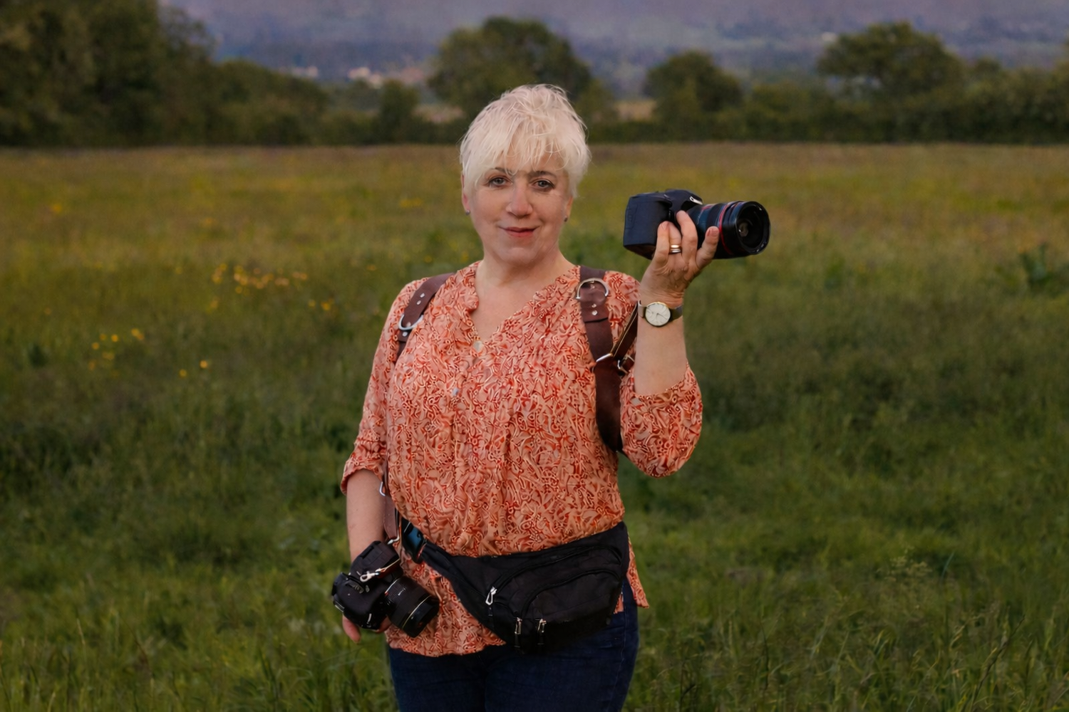 A photographer in a peach-colored lace top stands in a grassy meadow with camera equipment at golden hour.
