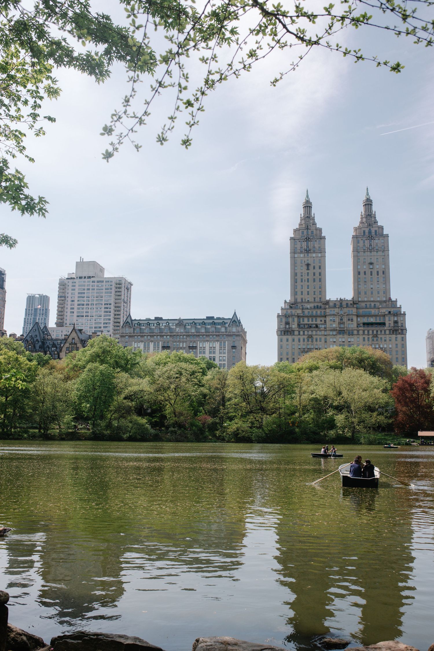 People rowing boats on Central Park Lake with iconic Upper West Side buildings and The San Remo towers in the background.