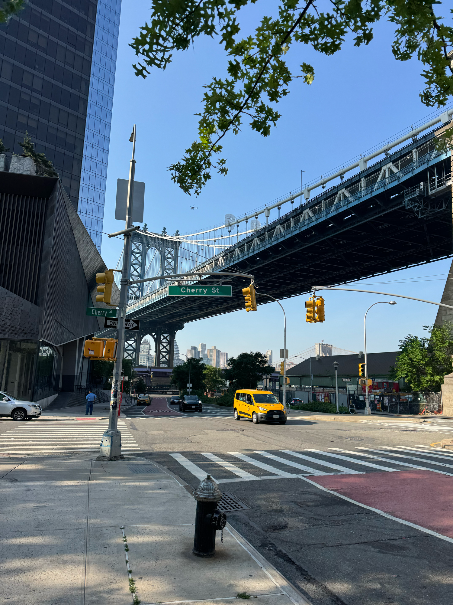 View of Manhattan Bridge from street level with yellow taxi passing underneath on a sunny day in New York City.