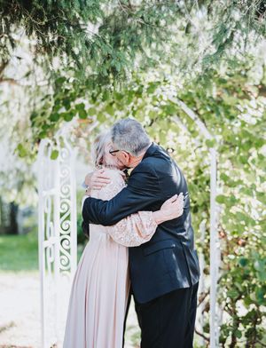 Couple embracing in a romantic hug under a white garden arch surrounded by lush greenery at an outdoor wedding.