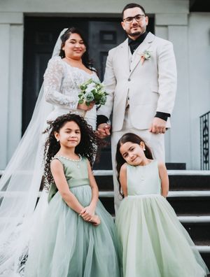 Bride and groom in white suit pose with two flower girls in sage green dresses on church steps.