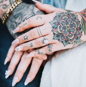 Two tattooed hands clasped together, one wearing a diamond ring, with intricate mandala and small tattoo designs.