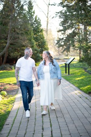 Couple holding hands walking on brick path in sunny park, man with tattoos in white tee, woman in white dress and denim jacket.