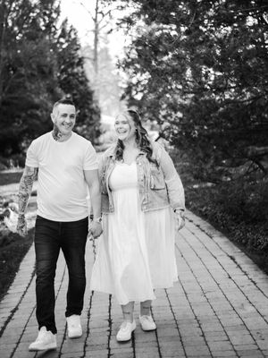 Black and white photo of a smiling couple walking hand in hand on a brick path surrounded by trees.