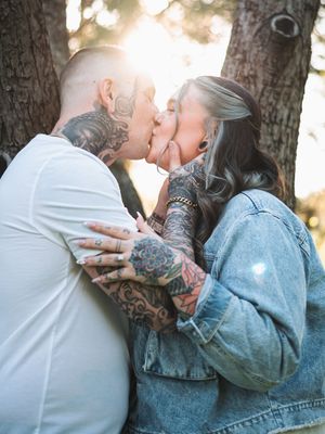 Couple with tattoos sharing a romantic kiss outdoors among pine trees with golden backlight.