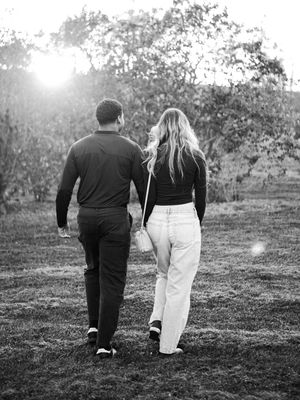 Black and white photo of a couple walking hand in hand through a sunlit grassy field with trees in the background.