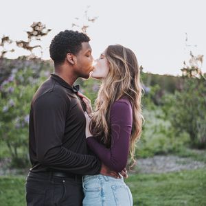 A couple shares a romantic kiss outdoors surrounded by lush greenery during golden hour.