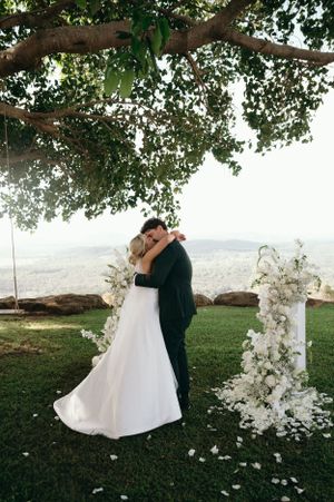 Couple embraces under a tree during outdoor wedding ceremony with floral arch.
