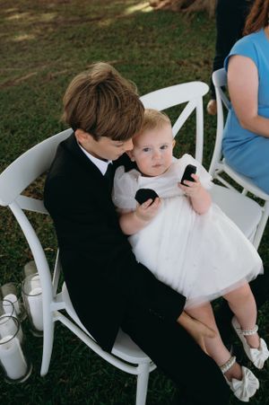 Young children in formal attire sit together on white chair at outdoor wedding.
