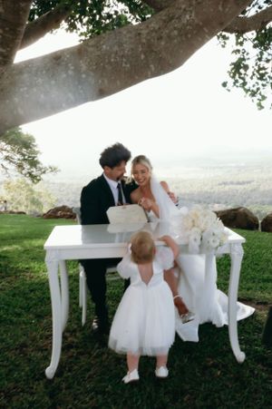 Wedding couple cuts cake at elegant outdoor reception table while young attendant watches.