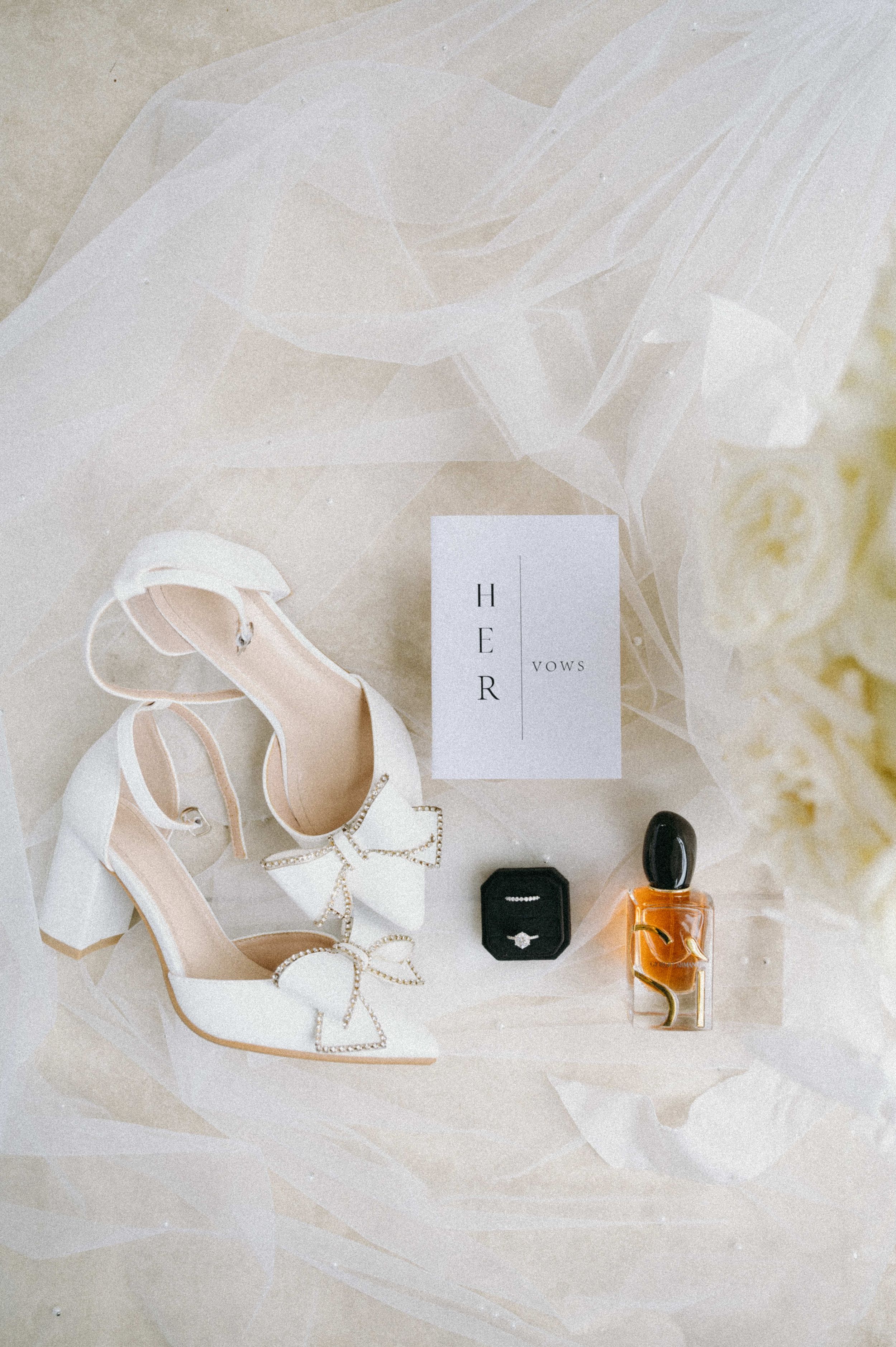 Wedding accessories flatlay with white heels, perfume bottle, and ring box on tulle fabric.