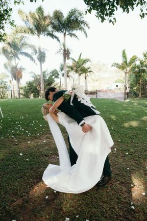 Romantic dip kiss between newlyweds on grassy lawn with palm trees.