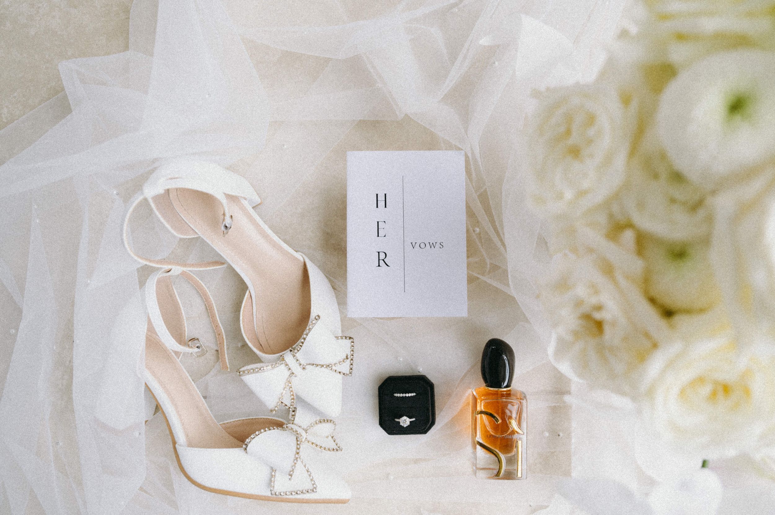 Bridal details including white bow heels, perfume, and jewelry box arranged on delicate tulle.