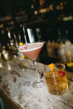 Two craft cocktails on a marble bar counter, one pink martini and one amber drink with orange garnish.