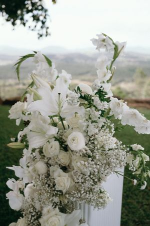 Elegant white floral arrangement with lilies, roses and baby's breath against a scenic outdoor backdrop.