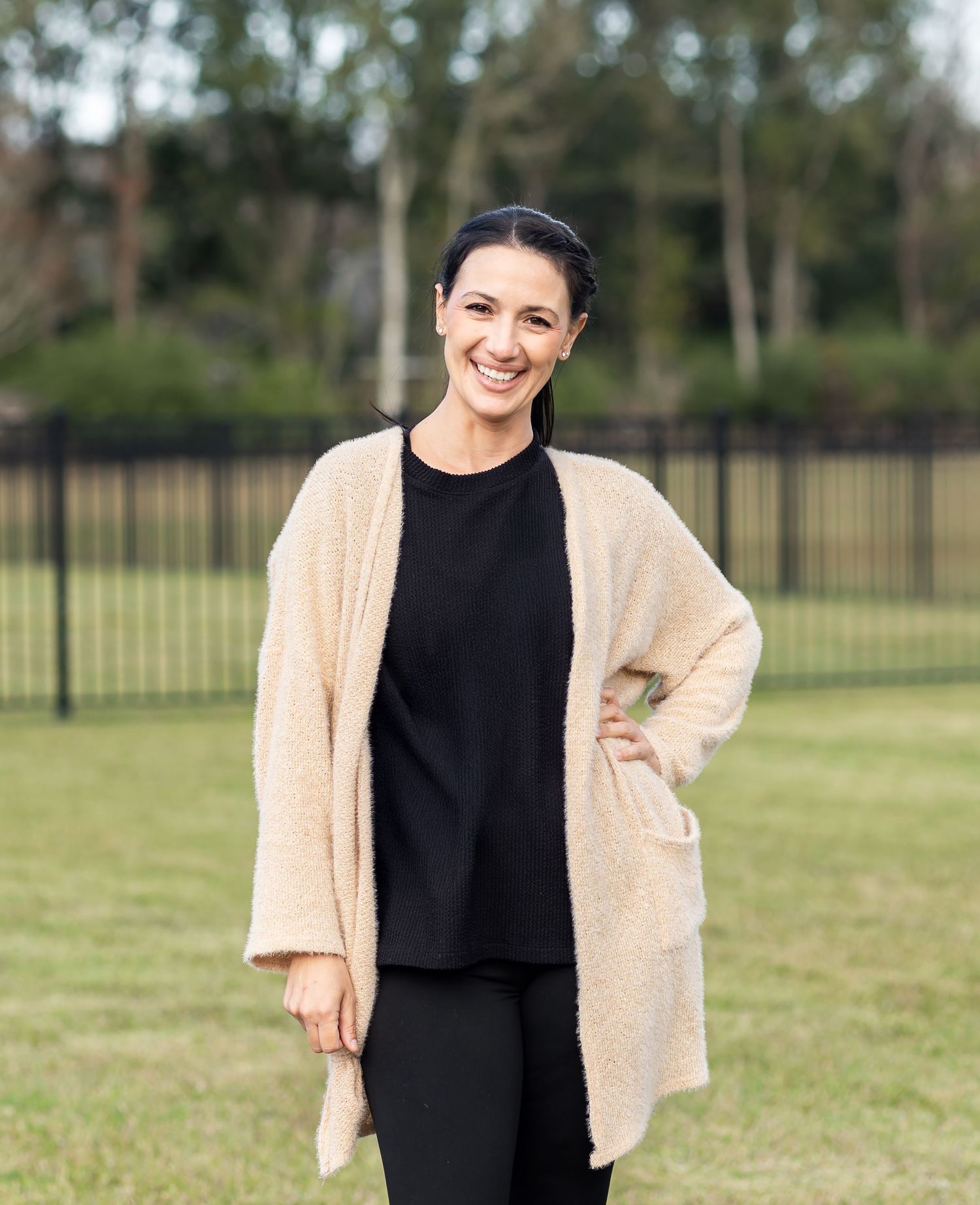 A person wearing a cozy beige cardigan and black top stands in a grassy field with trees in the background.