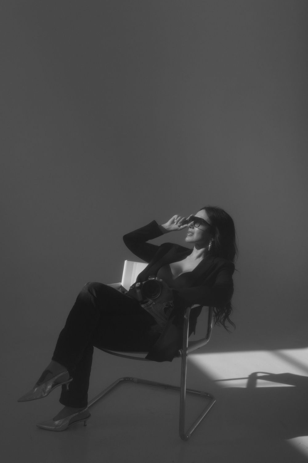 A person in dark clothing reclines dramatically in a chair against a gray background with stark lighting and shadows.
