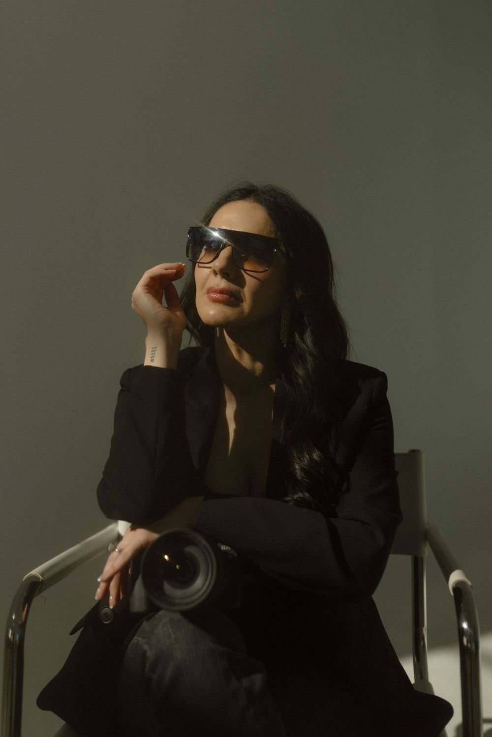 A person in a black outfit and sunglasses poses dramatically in a chair against a dark background.