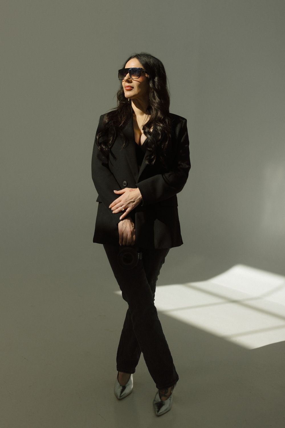 Stylish woman in black blazer and sunglasses poses confidently against grey wall with dramatic window lighting.