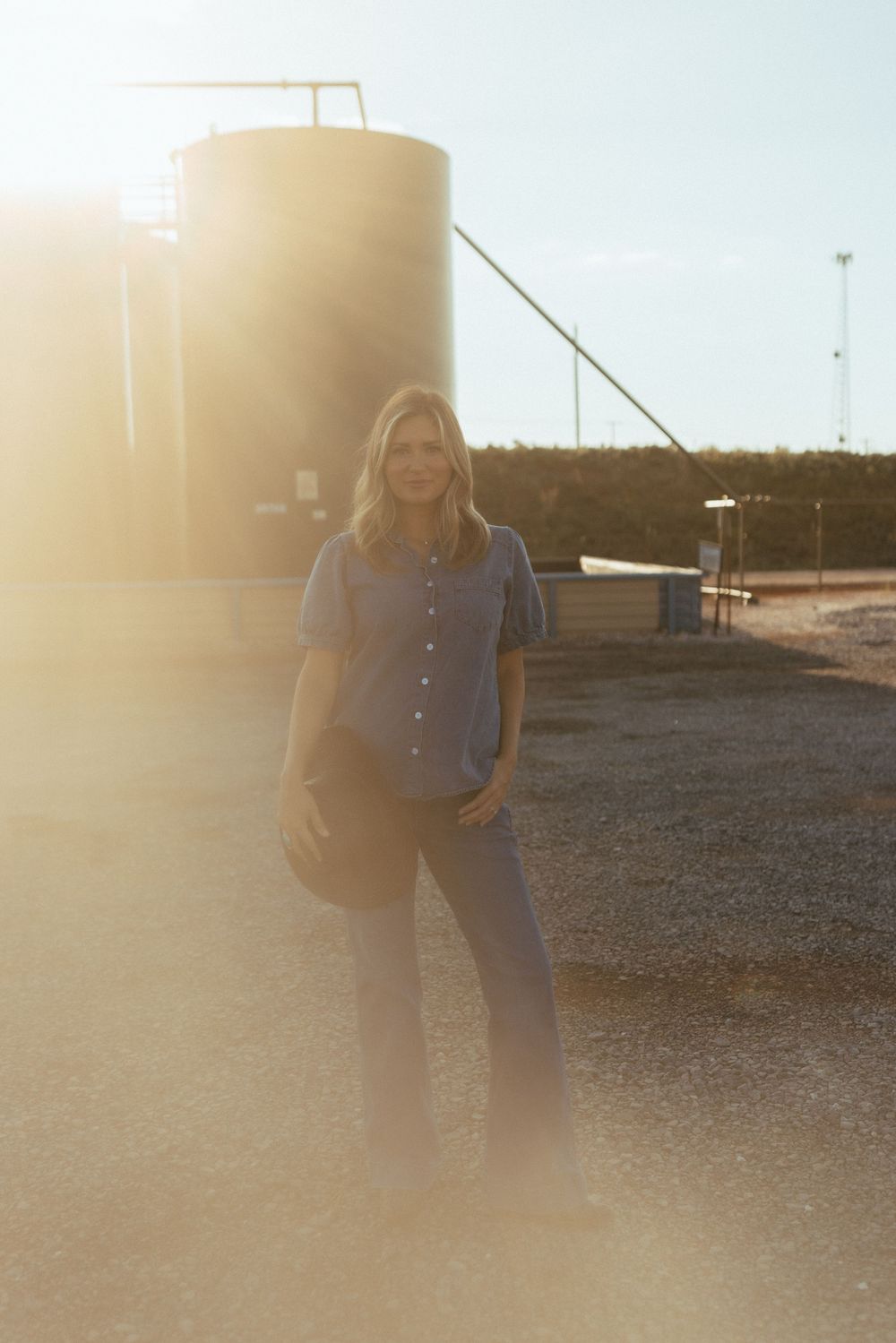A person stands in casual attire against an industrial backdrop with storage tanks at sunset creating a warm backlit glow.