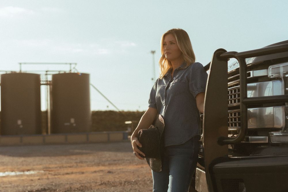A person in casual attire stands beside a livestock trailer during golden hour at a rural farm with storage tanks in background.