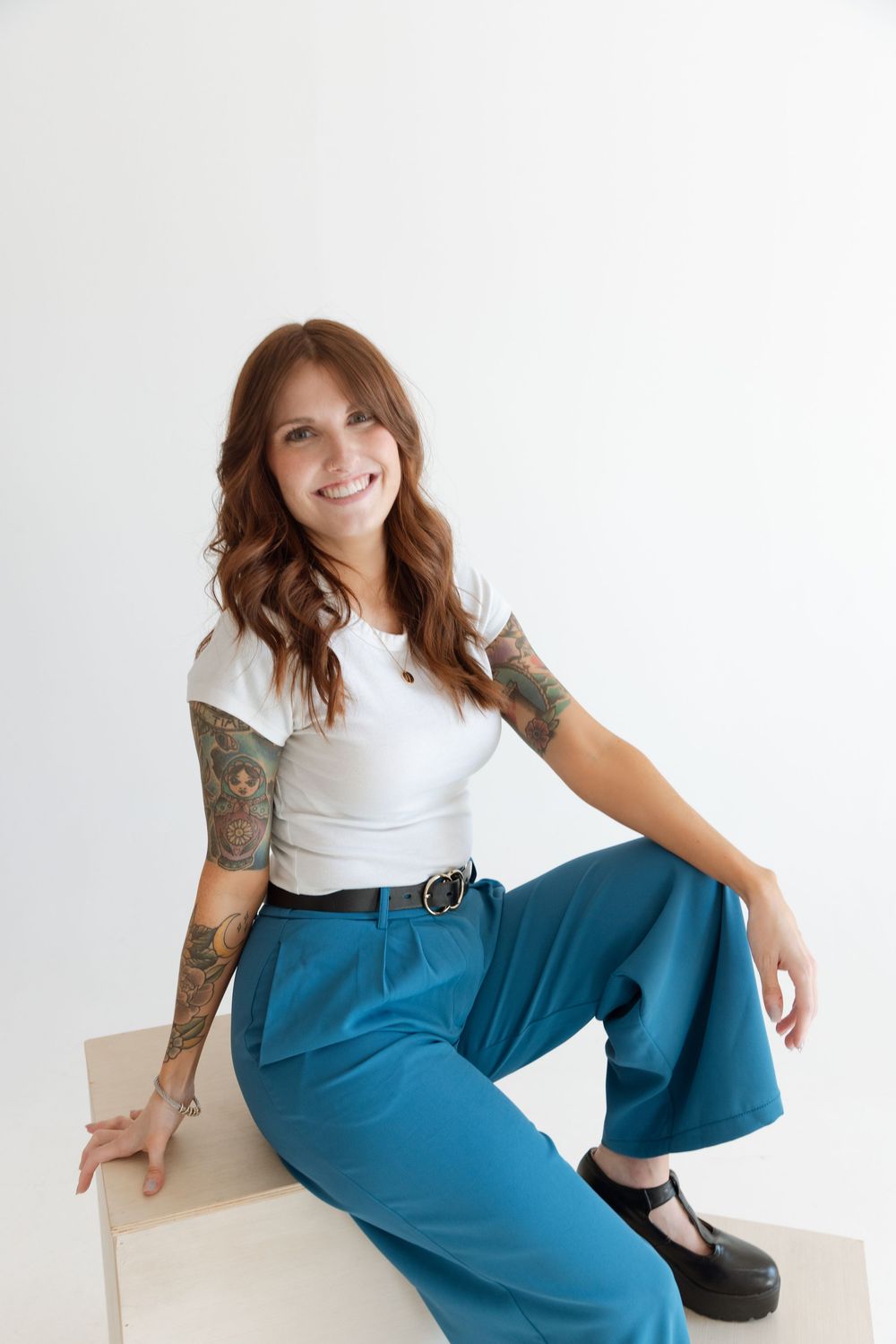 A person in a white t-shirt and teal wide-leg pants sits casually on a white surface, showing off colorful arm tattoos.