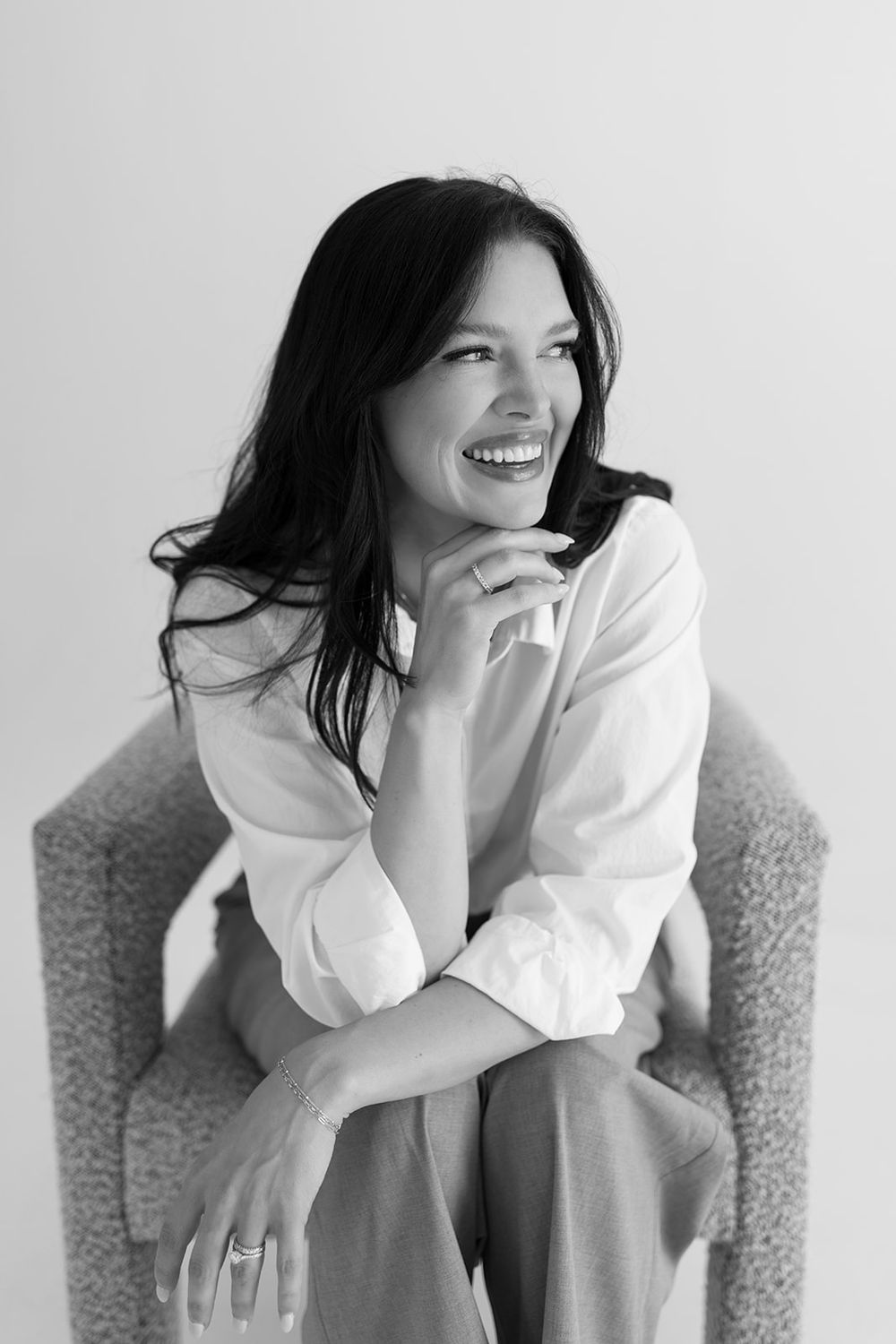Black and white portrait of someone with dark hair wearing a white shirt, sitting and smiling warmly in a chair.
