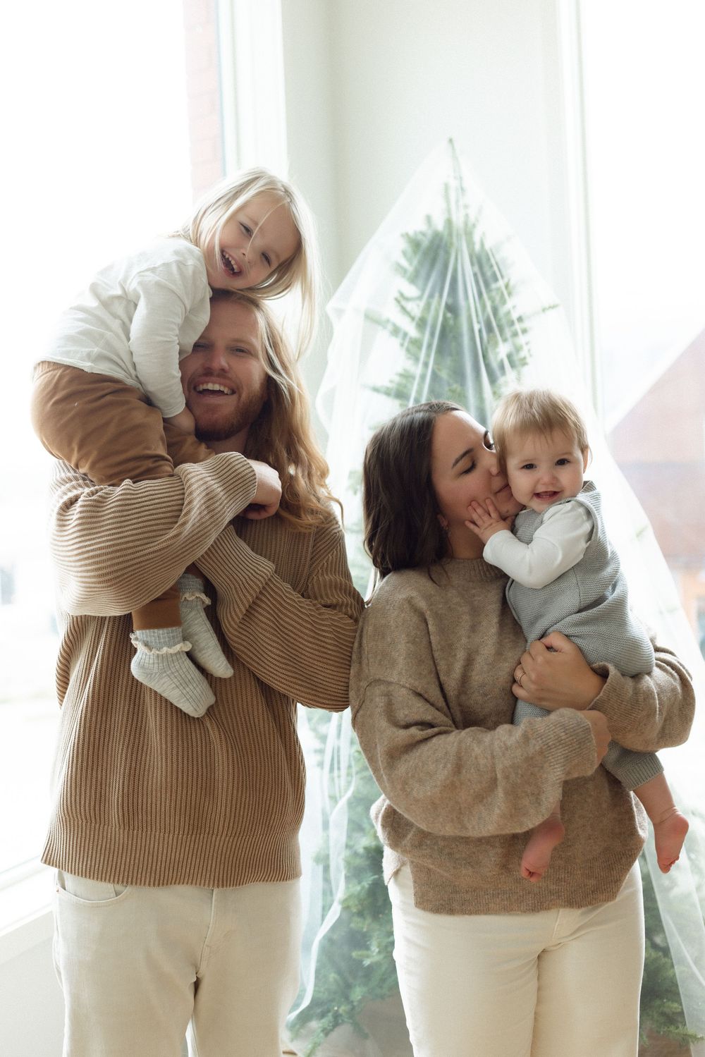 A family in matching beige sweaters and white pants poses together by a Christmas tree in a bright interior.