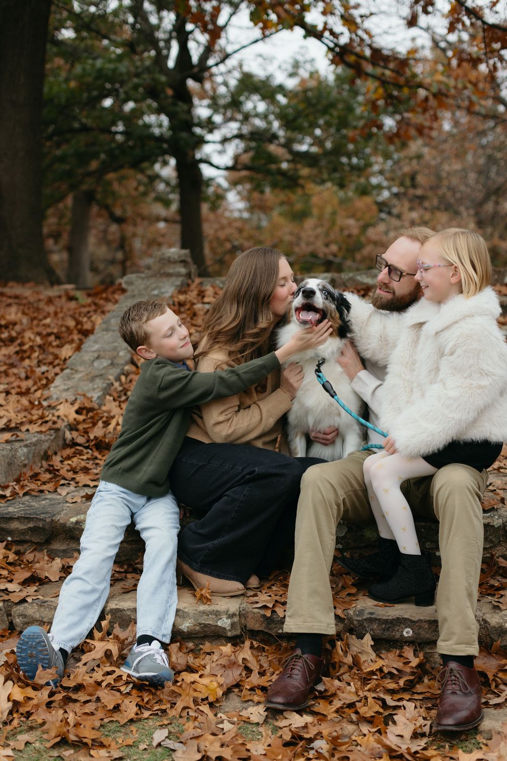 A family sits together on stone steps covered in autumn leaves while playing with their small dog.